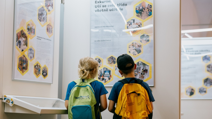Dvě malé děti s batohy si prohlížejí informační panely na výstavě, kterou pro veřejnost pořádá centrum robotiky.
