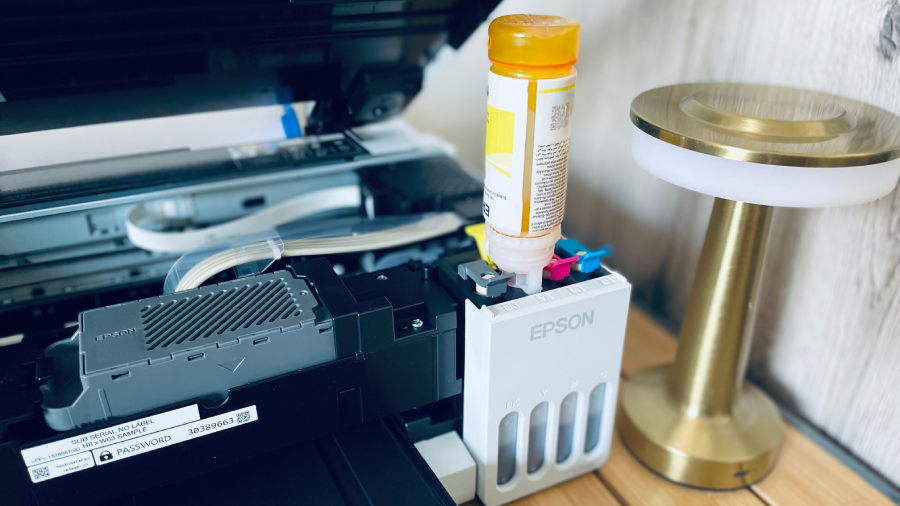 Pohled na doplňování žlutého inkoustu do integrovaných nádržek tiskárny Epson EcoTank L3366. Originální lahvička je otočená dnem vzhůru a zajištěná ve specifickém plnicím otvoru. Na přední straně tiskárny jsou vidět průhledové indikátory hladiny inkoustu a vedle přístroje stojí moderní zlatá stolní lampa.