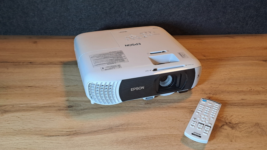 Projektor Epson EB-FH18 na dřevěném stole s dálkovým ovládáním položeným vedle něj.
