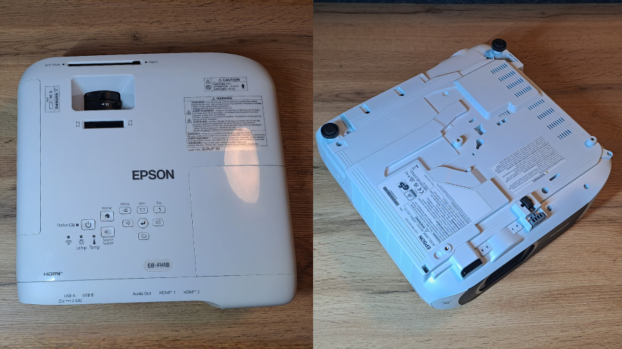 Horní a dolní pohled na bílý projektor Epson EB-FH18 umístěný na dřevěném stole, zobrazující ovládací panel, tlačítka pro vyhledávání zdroje, stavové kontrolky a označení vstupů.