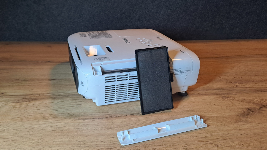 Projektor Epson EB-FH18 s vyjmutým vzduchovým filtrem a jeho plastovým krytem položenými před přístrojem na dřevěném stole.