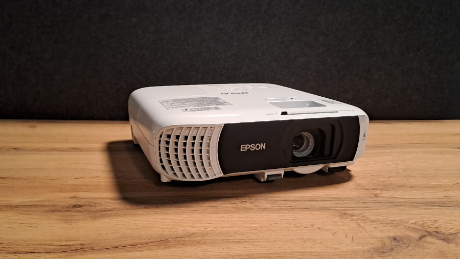Přední pohled na projektor Epson EB-FH18 s čočkou objektivu, ventilační mřížkou a nápisem Epson, umístěný na dřevěném povrchu před tmavě šedým pozadím.