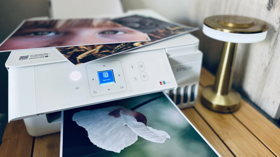 Tiskárna Epson EcoTank L3366 tiskne velkoformátovou fotografii A4 a na její horní části jsou položené hotové vytištěné fotky.