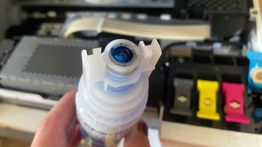 Detailní pohled na speciální bajonetový ventil lahvičky s modrým inkoustem Epson EcoTank se zámkovým mechanismem proti rozlití, držené v ruce nad otevřenými tankovými zásobníky tiskárny.