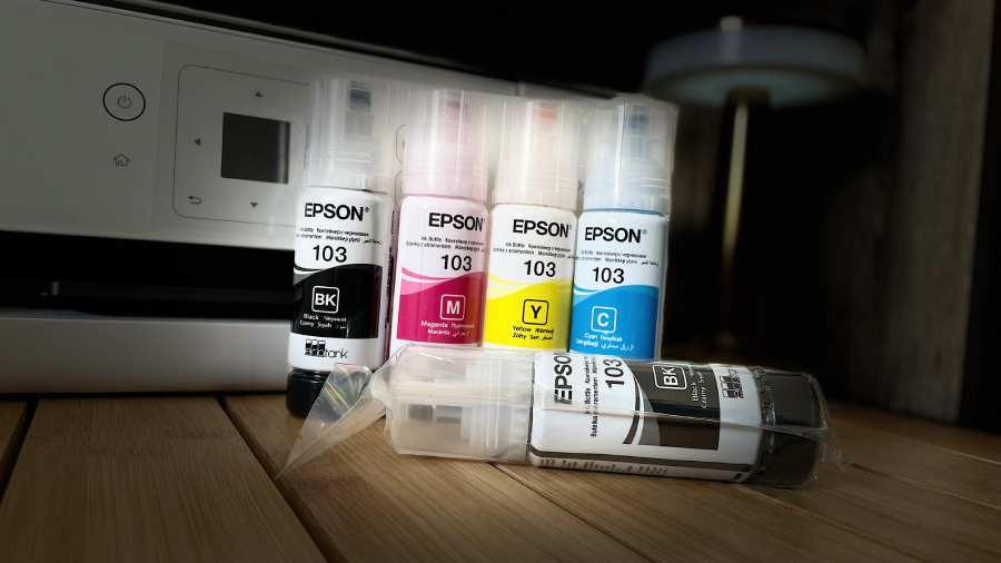 Sada čtyř originálních lahviček s inkoustem Epson 103 EcoTank (černá, azurová, purpurová, žlutá) stojících před bílou tiskárnou, s pátou černou lahvičkou v plastovém obalu ležící v popředí.