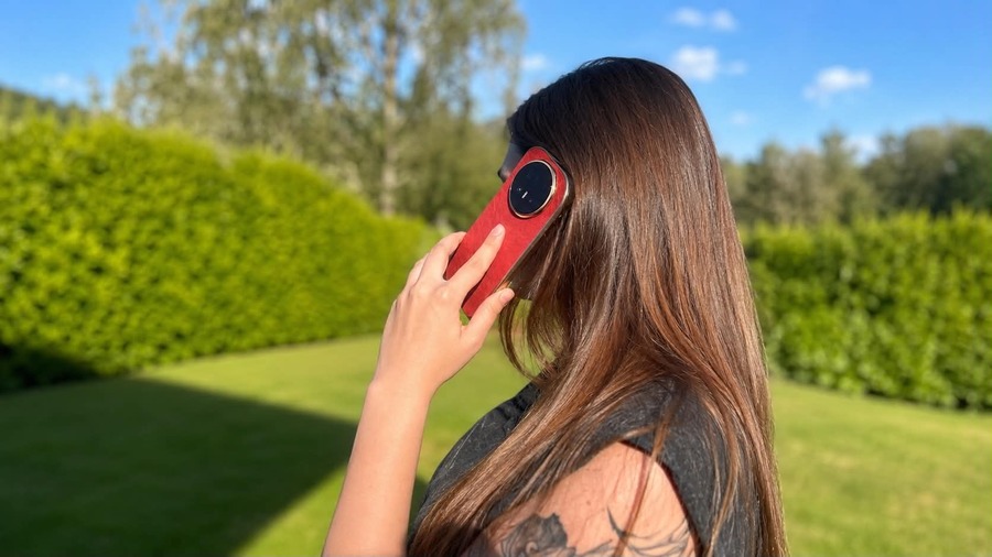Žena s dlouhými hnědými vlasy držící složený červený telefon HONOR Magic V6 u ucha při telefonování venku v zahradě.