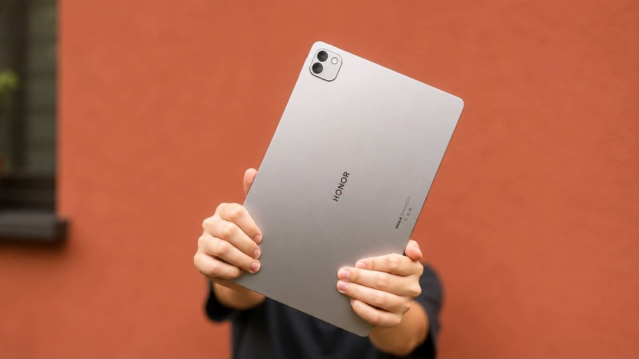Detailní pohled na zadní stranu tabletu HONOR MagicPad 4 s logem a fotoaparátem, který drží člověk před sebou proti rozostřené červené stěně.