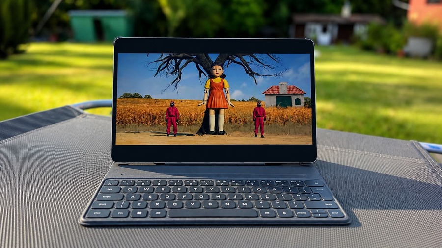 Tablet HONOR MagicPad 4 s připojenou klávesnicí stojí na venkovním lehátku, na displeji je scéna ze seriálu "Squid Game" s panenkou a dvěma strážci.
