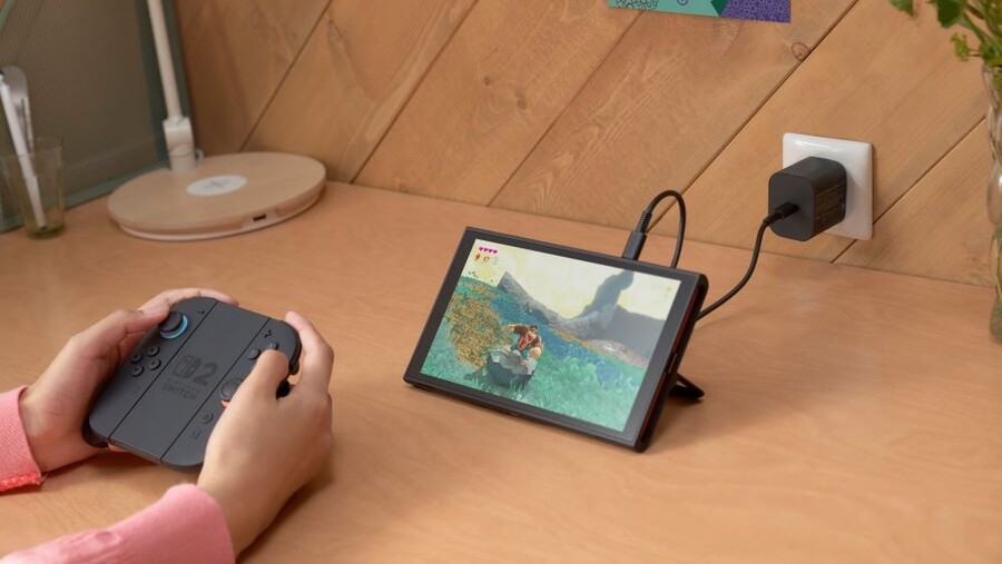 Člověk hrající na herní konzoli Nintendo Switch 2 v kapesním režimu, zatímco je zařízení připojeno k nabíječce na dřevěném stole.