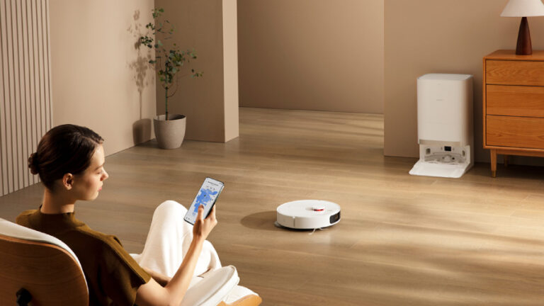 Bílý robotický vysavač Xiaomi Robot Vacuum X20+ uklízí dřevěnou podlahu v moderním interiéru, zatímco žena v popředí sleduje na mobilním telefonu digitální mapu úklidu domácnosti.