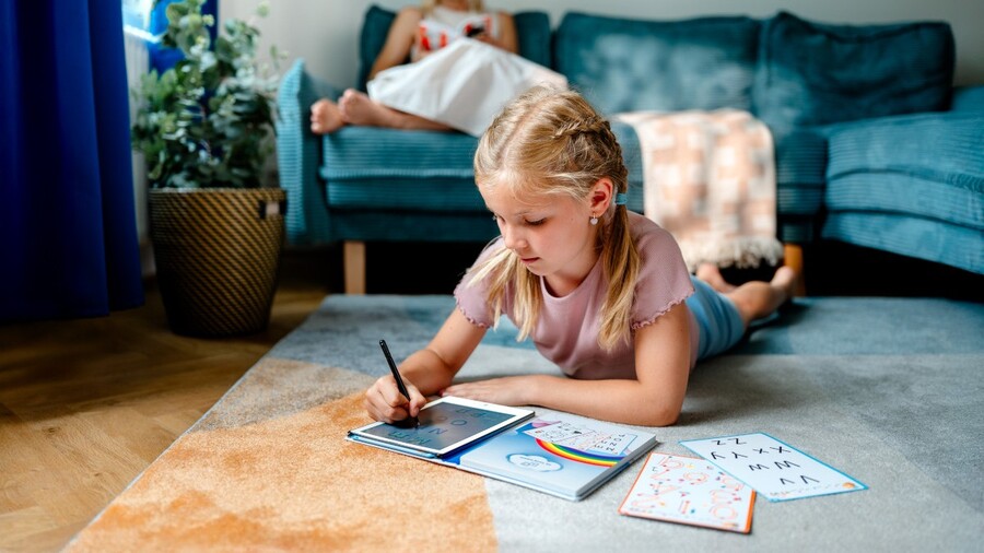 Back to school: Dívka leží na koberci a kreslí stylusem na tablet značky LAMAX PencilKid1, přičemž před sebou má předlohy.