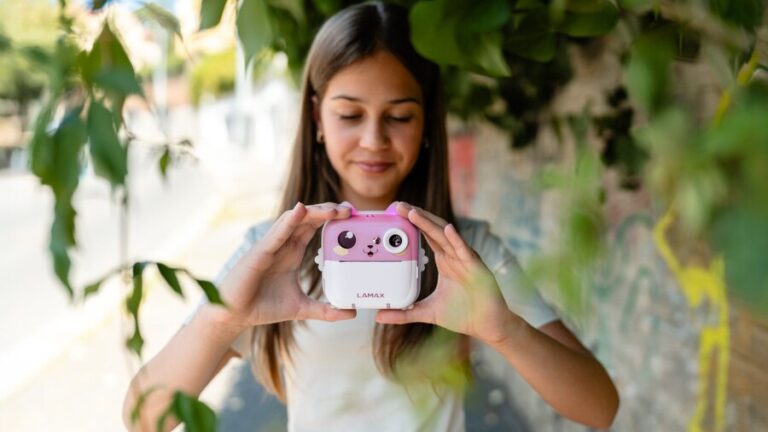 Dívka drží před sebou růžový dětský instantní fotoaparát LAMAX InstaKid2 AI Plus s motivem kočičky.