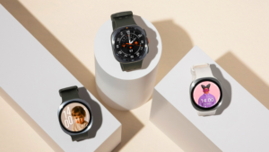Studiové aranžmá tří chytrých hodinek Samsung: uprostřed tmavé Galaxy Watch Ultra2 na bílém válci, vlevo dole tmavé Galaxy Watch9 s ciferníkem s fotografií dítěte a vpravo dole světlé Galaxy Watch9 s ciferníkem s kreslenou postavou.