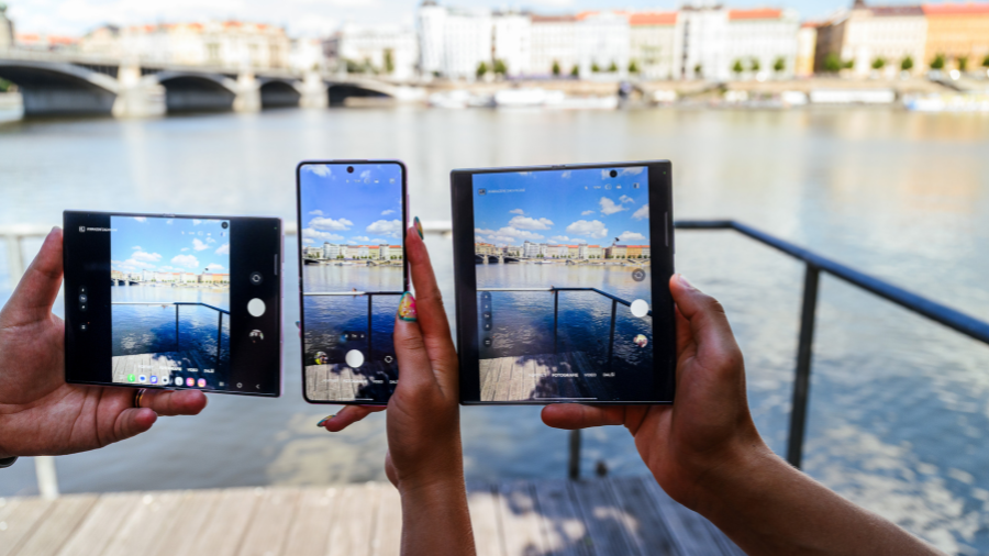 Tři lidé drží venku u vody různá zařízení řady Samsung Galaxy Z se zapnutými fotoaparáty, které zachycují okolní krajinu s řekou a mostem.