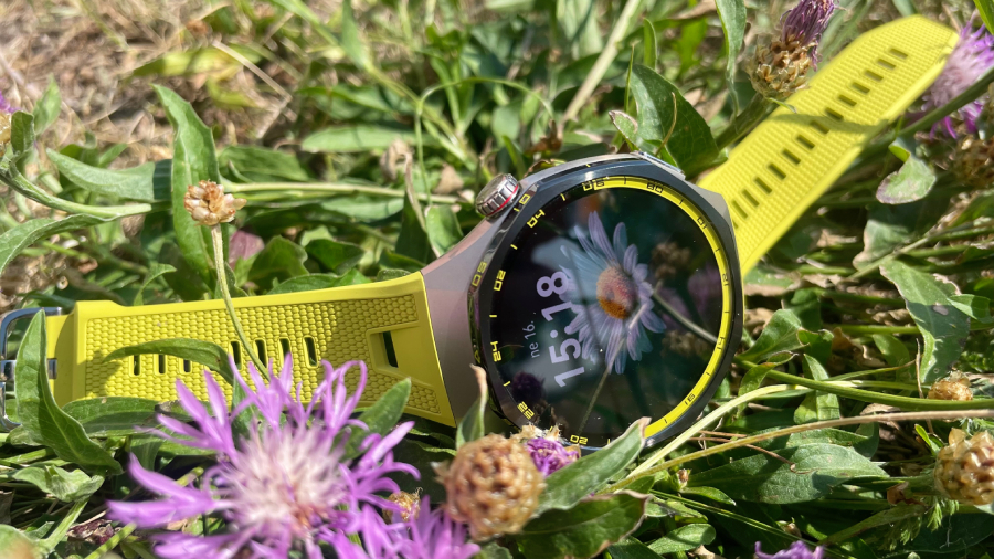Chytré hodinky Huawei Watch GT 7 Pro s olivovým sportovním řemínkem položené v trávě mezi rozkvetlými fialovými květy jetelíčku.