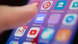 Detailní pohled na prst dotýkající se obrazovky chytrého telefonu, kde svítí ikona aplikace Myspace obklopená dalšími platformami jako Gmail, YouTube nebo Pinterest.