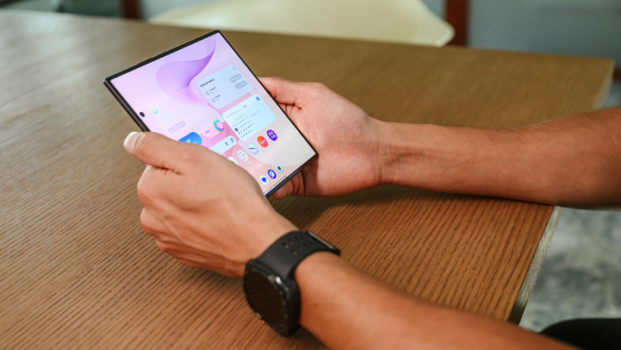 Pohled na ruce držící rozložený ohebný displej chytrého telefonu Samsung Galaxy Z Fold8 Ultra nad dřevěným stolem s rozsvícenou domovskou obrazovkou a widgety.