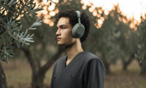 Mladý muž s tmavými kudrnatými vlasy pózuje z profilu otočený doleva a má nasazená nová bezdrátová sluchátka Sony WH-1000XM6 v olivově zelené barvě. Stojí uprostřed olivového háje obklopený stromy a listy, přičemž v pozadí prosvítá jemné světlo zapadajícího slunce. Muž má na sobě elegantní, minimalistický šedý oděv bez límečku, který ladí s přírodním a klidným tónem celé scény.