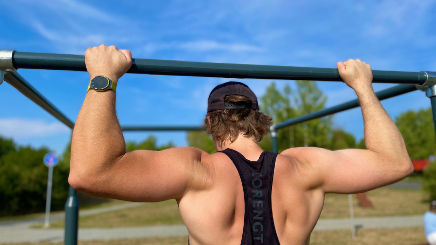 Zadní pohled na muže provádějícího přítahy na tmavé workoutové konstrukci pod širým nebem, který má na levém zápěstí hodinky Huawei Watch GT 7 Pro s olivovým řemínkem.