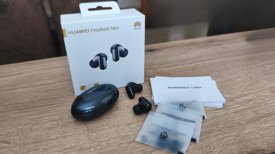 Balení sluchátek Huawei FreeBuds Neo s krabičkou, nabíjecím pouzdrem, sluchátky, náhradními silikonovými špunty a záručním listem položenými na dřevěném stole.
