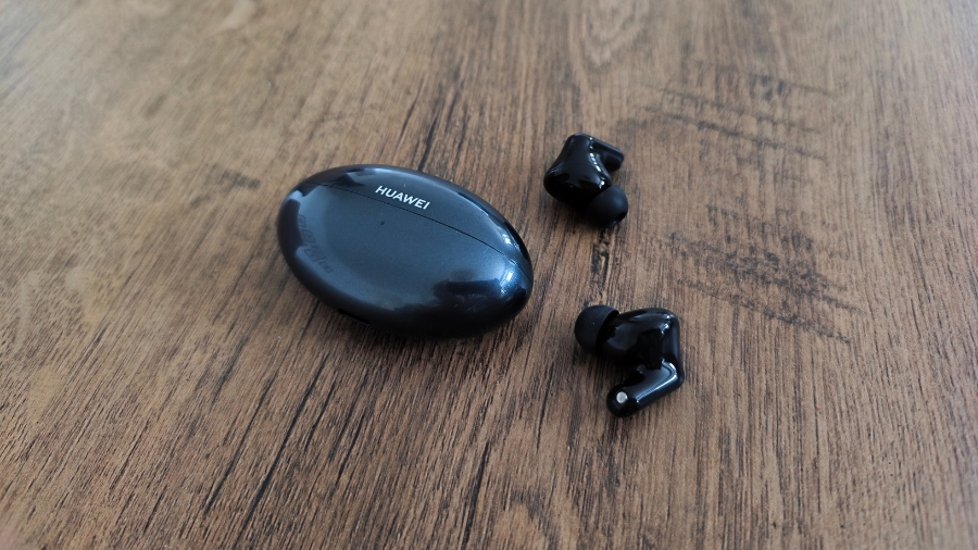 Černé bezdrátové sluchátka Huawei FreeBuds Neo položená vedle zavřeného nabíjecího pouzdra na dřevěném povrchu.