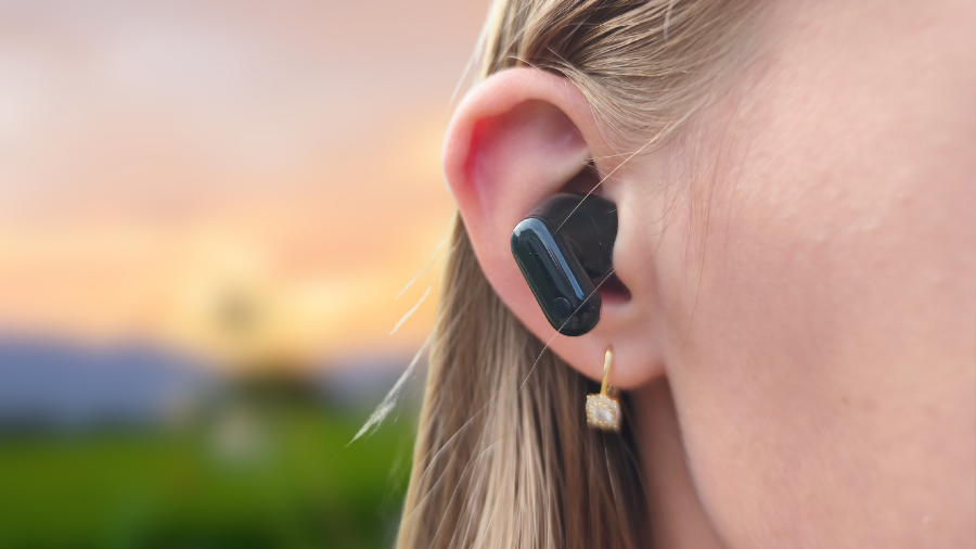 Detailní pohled na ženu s černým bezdrátovým sluchátkem Huawei FreeBuds Neo v uchu na rozostřeném přírodním pozadí.