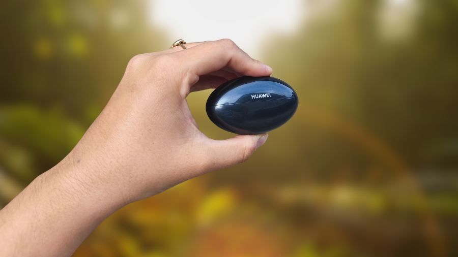 Detailní pohled na ruku držící zavřené černé pouzdro sluchátek Huawei FreeBuds Neo na rozostřeném podzimním pozadí.