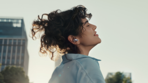 Žena se zavřenýma očima v městském prostředí nosí stříbrné bezdrátové sluchátko.