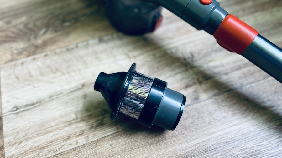 Vyjmutý omyvatelný cyklónový filtr z tyčového vysavače JIGOO C200 Cordless Vacuum Cleaner položený na vinylové podlaze vedle sací trubice.