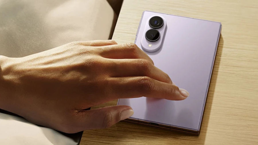 Ruka natahující se z postele k levandulovému telefonu Samsung Galaxy Z Fold8, který leží složený na dřevěném nočním stolku a ukazuje detail zadního fotoaparátu.