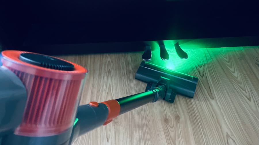 Pohled na tyčový vysavač JIGOO C200 Cordless Vacuum Cleaner se zapnutým zeleným LED osvětlením podlahové hubice při vysávání pod pedály klavíru.