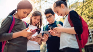 Čtyři náctiletí školáci s batohy na zádech stojí venku v kruhu a soustředěně sledují displeje svých chytrých telefonů, což ilustruje téma kyberšikana na školách.