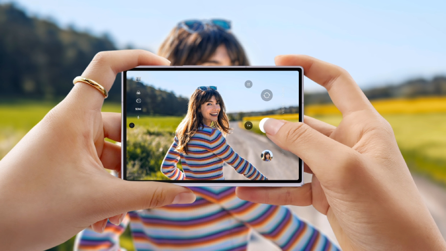 Pohled na rozložený displej telefonu Samsung Galaxy Z Fold8 v rukou uživatele, který fotografuje usmívající se ženu v pruhovaném svetru na venkovní cestě.
