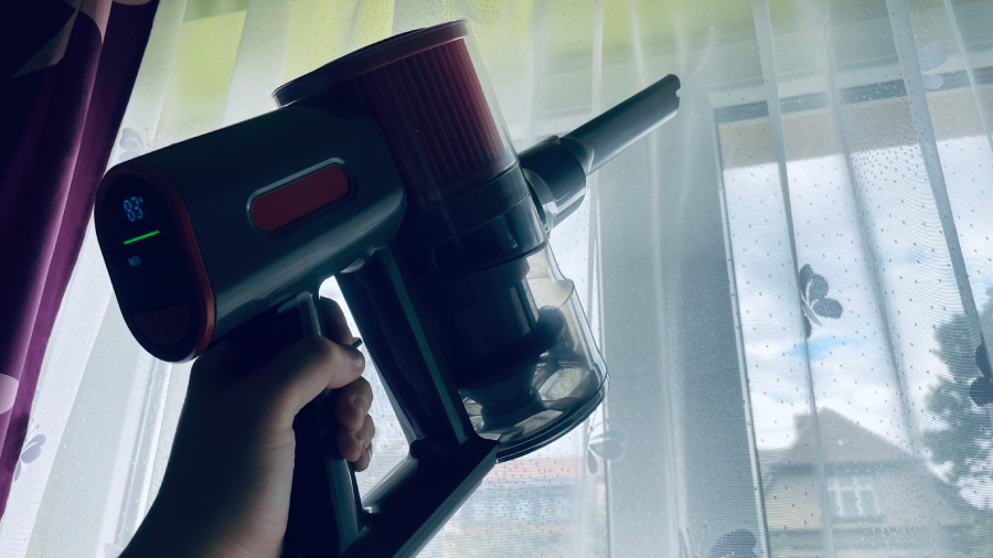 Detailní pohled na ruční jednotku vysavače JIGOO C200 Cordless Vacuum Cleaner se štěrbinovou hubicí při vysávání prachu ze záclon u okna.