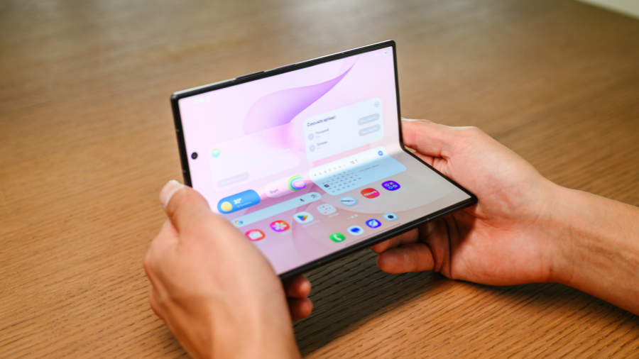 Detailní pohled na lidské ruce držící částečně rozevřený skládací smartphone Samsung s rozsvíceným vnitřním displejem a widgety na dřevěném stolu.