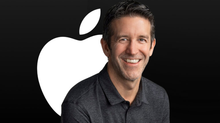 Portrét usmívajícího se Johna Ternuse, generálního ředitele společnosti Apple, v tmavě šedé polokošili, za nímž se na černém pozadí nachází velké bílé logo firmy.
